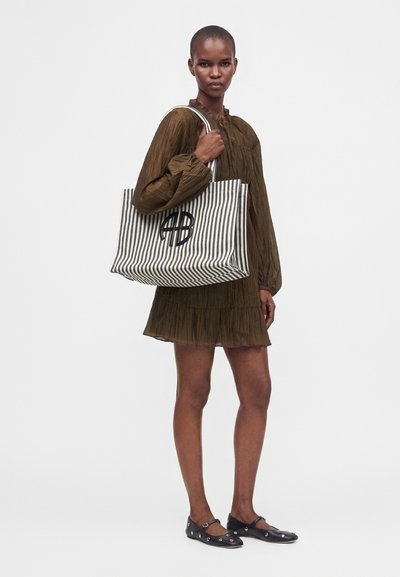Femme en robe marron à manches longues tenant un grand sac cabas rayé noir et blanc avec un logo noir, portant des chaussures plates noires.