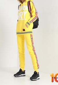 Geel hoodie met witte "SUPERDRY" tekst en zwarte accenten, gecombineerd met bijpassende joggers met zijkant in rood en witte branding. Zwarte sneakers.