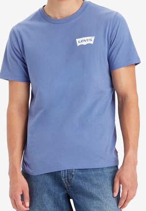 Homme portant un t-shirt Levi's bleu clair et un jean bleu, montré du cou aux hanches sur fond blanc.