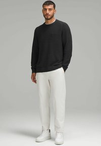 Schwarzer Strickpullover mit rundem Ausschnitt, strukturiertem Stoff, kombiniert mit weißen Hosen und weißen Sneakers. Das Modell steht vor einem neutralen Hintergrund.
