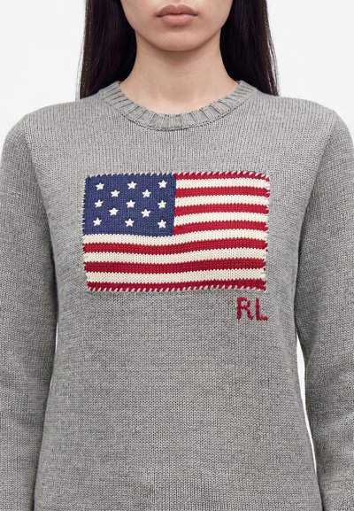 Jeune personne portant un pull en maille gris avec un drapeau américain brodé et les initiales rouges "RL" sur la poitrine.
