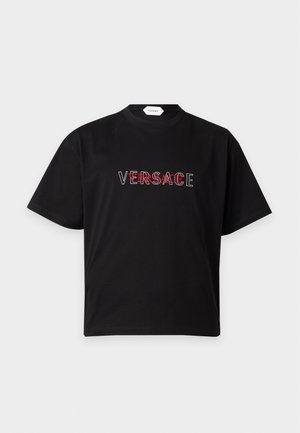 Mężczyzna w czarnej koszulce Versace, kremowych spodniach, białych i brązowych sneakersach oraz czarnych okularach przeciwsłonecznych stoi z rękami w kieszeniach.