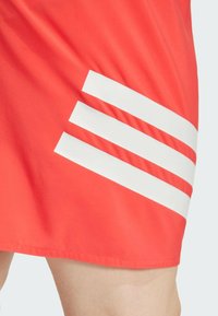 Pantalones cortos deportivos rojos hechos de tela ligera, con tres franjas diagonales blancas en la parte inferior izquierda y un dobladillo cosido.