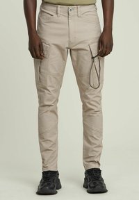 Pantaloni cargo beige con multiple tasche, dettaglio con cordoncino, design affusolato e texture liscia, abbinati a scarpe sportive nere.