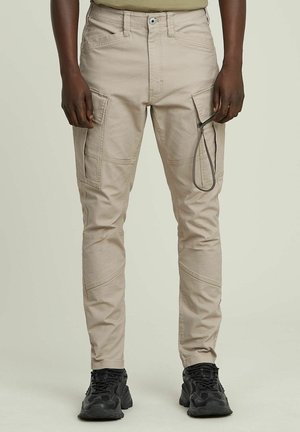 Beige cargo pants with multiple pockets, a drawstring accent, tapered design, and a smooth texture, paired with black athletic shoes.