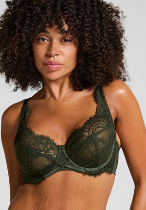 Vrouw met krullend haar, gekleed in een donkergroene kanten bh, weergegeven van schouders tot midden torso tegen een effen grijze achtergrond.