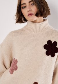 Crèmekleurige gebreide trui met een hoge ribbelscol, versierd met overgrote bloemenapplicaties in donker bordeaux en lichtroze aan de voorkant.