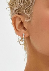 Orecchini d'argento con design a goccia e cristalli trasparenti, sospesi da una piccola ovale superiore. Lo sfondo è neutro.