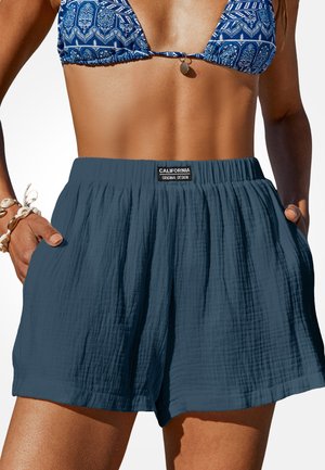 Frau trägt blau gemustertes Bikinioberteil und lockere dunkelblaue strukturierte Shorts mit dem Aufnäher "California Original Design", Hände in den Taschen.