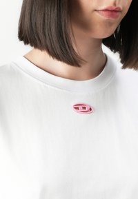 Nærbilde av en person som har på seg en hvit skjorte med en liten rosa oval brodert logo nær kragen og rett mørkebrunt hår.