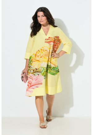 Vrouw draagt een gele V-halsjurk met grote multicolor bloemenpatronen, houdt een roze clutch vast en draagt beige sandalen met versieringen.