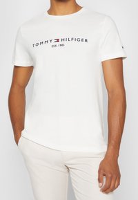 Bílý bavlněný tričko s kulatým výstřihem a krátkými rukávy. Logo "TOMMY HILFIGER EST. 1985" v námořnickém modrém s červenými a modrými akcenty. Hladká textura.