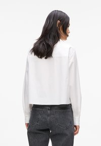 Hvit cropped skjorte med lange ermer, knappede mansjetter, glatt tekstur og en bakre yoke-detalj, kombinert med mørk grå høy midje jeans.