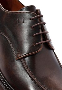 Zapato de vestir de cuero marrón con una textura suave, costuras mínimas y cordones marrones. Presenta una punta redondeada y un área de cordones expuesta.