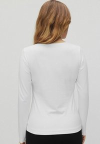 Weißes Langarmshirt mit einem runden Ausschnitt, aus glattem Stoff gefertigt. Es hat eine figurbetonte Silhouette und ein minimalistisches Design, keine sichtbaren Muster.