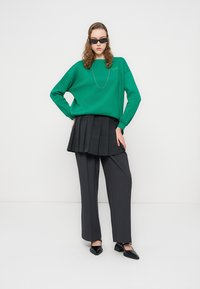 Πράσινο φαρδύ φούτερ με ribbed μανσέτες, συνδυασμένο με μαύρη πλισέ φούστα πάνω από γκρι παντελόνια και μαύρα μοκασίνια. Αξεσουάρ περιλαμβάνουν γυαλιά ηλίου.