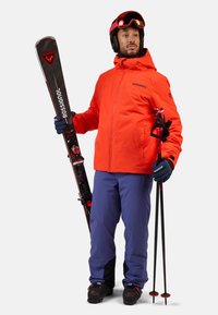Rode skijas, blauwe skibroek, zwarte skischoenen; houdt zwarte ski's en stokken vast. Jas heeft een rits en logo, met handschoenen voor grip.