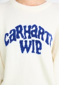 Kreemivärvi kudum, millel on tekstureeritud sinine "CARHARTT WIP" logo rinnal, ribidega ümar kaelus ja pehme kangas.