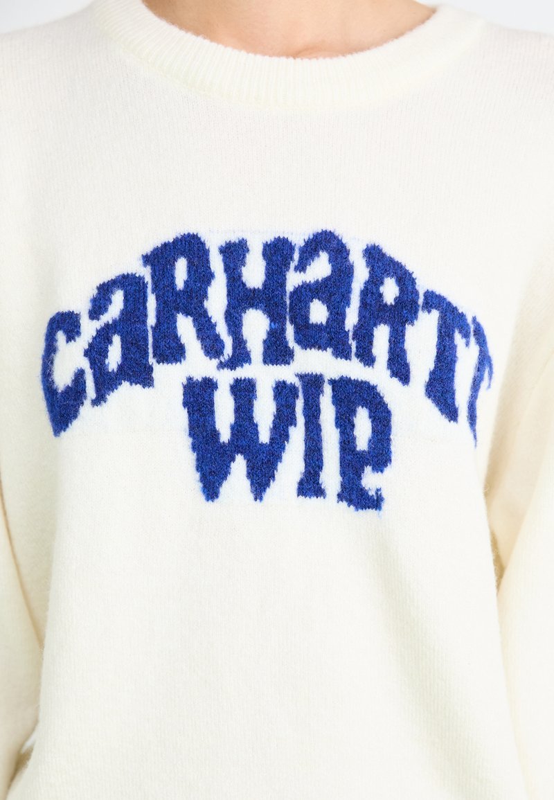 Crème-kleurige gebreide trui met een gestructureerd blauw "CARHARTT WIP" logo over de borst, voorzien van een geribbelde ronde hals en zachte stof.