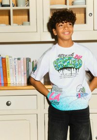 Weißes Grafik-T-Shirt mit bunten Designs und Text. Es hat kurze Ärmel und wird mit schwarzen Denim-Hosen kombiniert. Siebdruck-Akzente sichtbar.