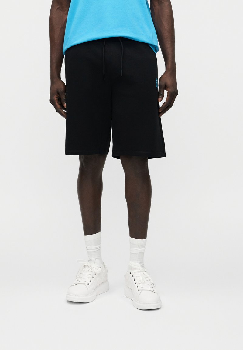 Άνδρας φορώντας μαύρα σορτς με κορδόνι, μπλε πουκάμισο, λευκές κάλτσες και λευκά αθλητικά παπούτσια, στέκεται μπροστά σε ένα απλό φωτεινό φόντο.