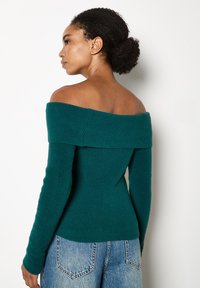 Donna con capelli ricci raccolti in un basso chignon, indossa un maglione a coste verde mare con spalle scoperte e jeans in denim azzurro chiaro, in posizione laterale contro un muro bianco.