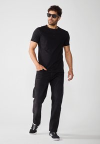Svart kortermet henley-skjorte, svarte jeans og sorte joggesko med hvite detaljer. Modell iført solbriller, går mot en lys bakgrunn.