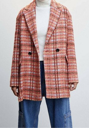 Vrouw draagt een rood-wit geruite dubbelgeknoopte jas over een witte coltrui en blauwe spijkerbroek met zakken.