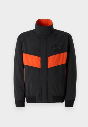 Zwarte jas met een oranje horizontale streep, hoge kraag, elastische manchetten en een logo patch op de borst. Soepele textuur en lichtgewicht materiaal.