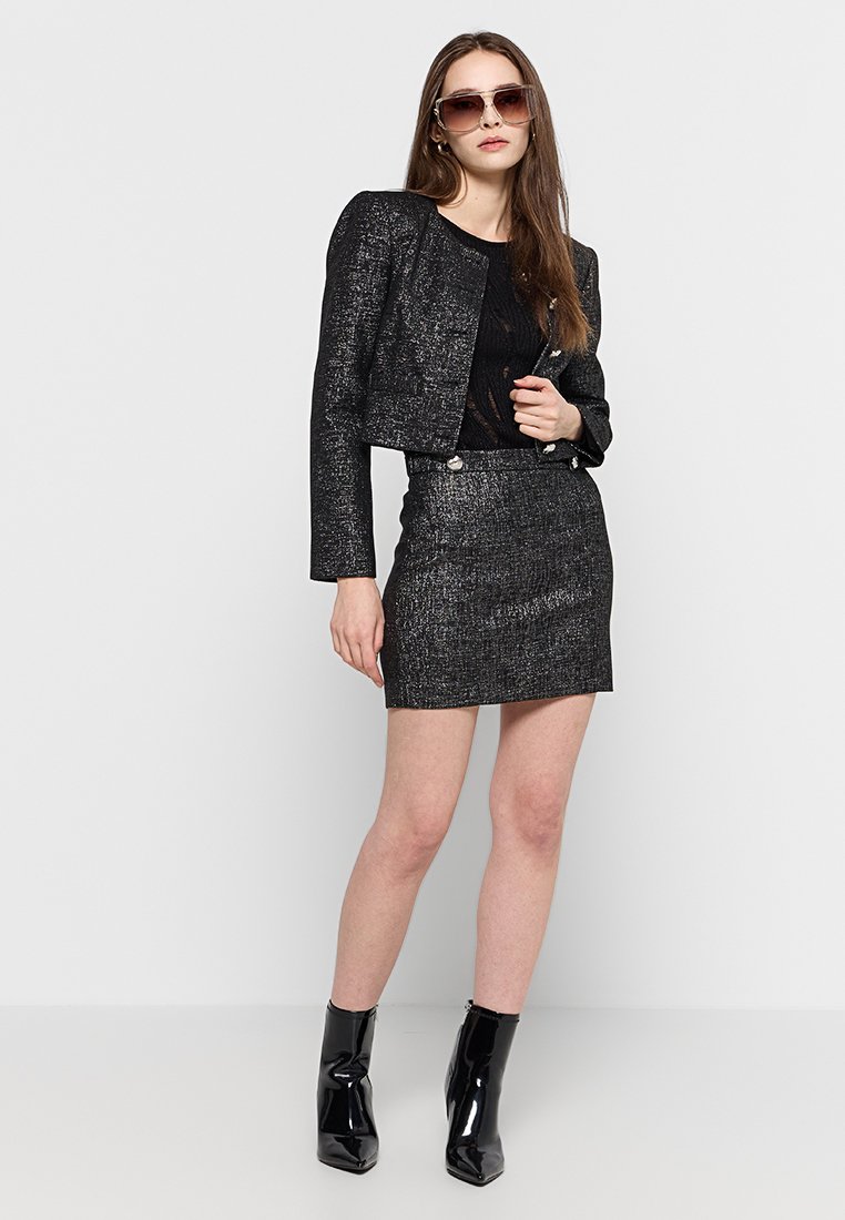 Veste noire cintrée et jupe ajustée avec un éclat métallique, associées à des bottines noires brillantes. Les accessoires comprennent des lunettes de soleil surdimensionnées.