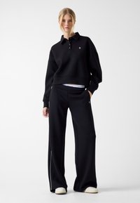 Czarna, oversize’owa bluza z kołnierzem i trzema guzikami, zestawiona z czarnymi spodniami o szerokich nogawkach z białymi paskami po bokach. Wykonana z miękkiej bawełnianej mieszanki.