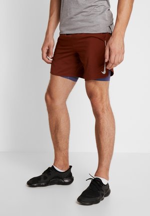 Mann trägt rostfarbene Sportshorts mit blauen inneren Kompressionsshorts, graues Tanktop und schwarze Laufschuhe, steht auf einem schlichten Boden.