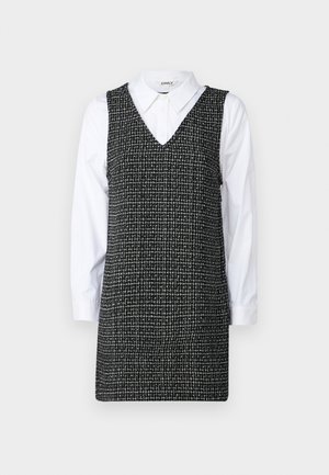 Rochie cu carouri alb-negru, cu decolteu în V și fără mâneci, stratificată peste o cămașă albă cu guler și mâneci lungi.