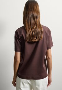 T-shirt di cotone marrone con maniche corte e scollatura rotonda. Tessuto morbido, vestibilità rilassata, mostrato da dietro, abbinato a pantaloni chiari.
