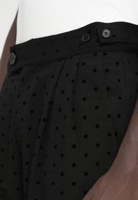 Calças pretas plissadas com padrão de bolinhas, apresentando dois botões pretos na cintura e uma textura de tecido suave.