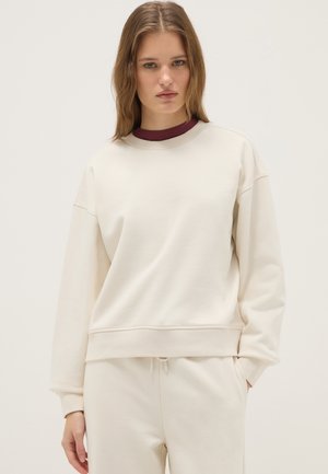 Junge Frau mit glattem hellbraunem Haar, die einen cremefarbenen, übergroßen Sweatshirt und passende Hosen auf neutralem Hintergrund trägt.