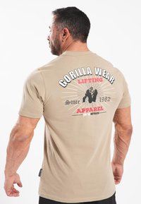 Šviesiai rudas medvilninis marškinėliai su grafiniu dizainu ant nugaros, kuriame yra išraiškingas tekstas ir gorilos iliustracija. Trumpomis rankovėmis, apvaliu kaklu.