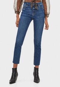 Jeans skinny azul oscuro con una cintura alta, detalles desgastados y un largo recortado. Combinados con botas negras hasta el tobillo.