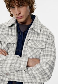 Harmaa-valkoinen ruudullinen takki pehmeästä kankaasta, jossa kaulus, nepparit ja kaksi rintataskua. Päällä farkkupaita.