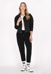 Jachetă neagră croită peste un tricou alb, asortată cu pantaloni sport negri și adidași albi. Modelul stă în fața unui fundal deschis.