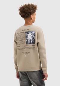 Tiener met krullend haar die een beige sweatshirt draagt met een stedelijk landschap en vliegende haaien grafiek en zwarte tekst op de achterkant.