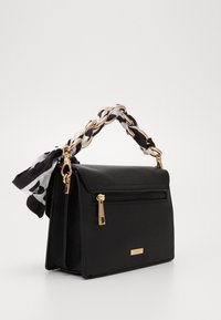 Bolso negro con una superficie texturizada, con un asa de cadena y una bufanda en blanco y negro atada. Detalles en dorado y cremallera visibles.