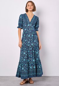 Maxikleid in Dunkelblau mit floralen Mustern in Türkis und Grün. Mit tiefem V-Ausschnitt, Puffärmeln und gestuften Rock. Braune Sandalen.