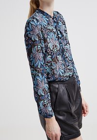 Långärmad blus i genomskinligt svart tyg med blå och lila blommönster, knappstängning på framsidan, rynkade ärmar, i kombination med svarta lädershorts.