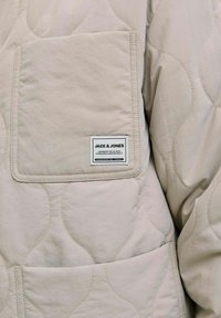 Veste matelassée beige avec une poche carrée. Elle présente une étiquette de marque et une texture lisse, mettant en valeur les motifs de couture et un matériau léger.