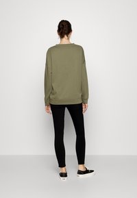 Oliivroheline ülelaia sweatshirt, millel on langenud õlad, koos mustade retuuside ja mustade tennistega, millel on valge tallaga ja ruudulised aktsendid.