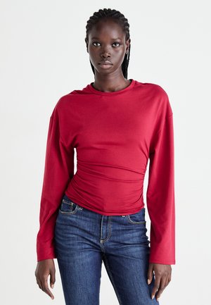 Rød langærmet top med samlet detalje i siden, rund halsudskæring og blødt stof, parret med blå tætsiddende jeans.