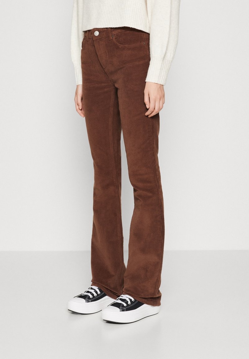 En person som bär bruna kordbyxor med knapp- och dragkedjestängning, tillsammans med svarta och vita platåsneakers och en krämfärgad tröja.