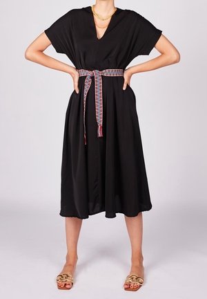 Sort kjole med V-udskæring, korte ærmer og en udskåret nederdel. Har et farverigt mønstret bælte og bæres med guld sandaler.