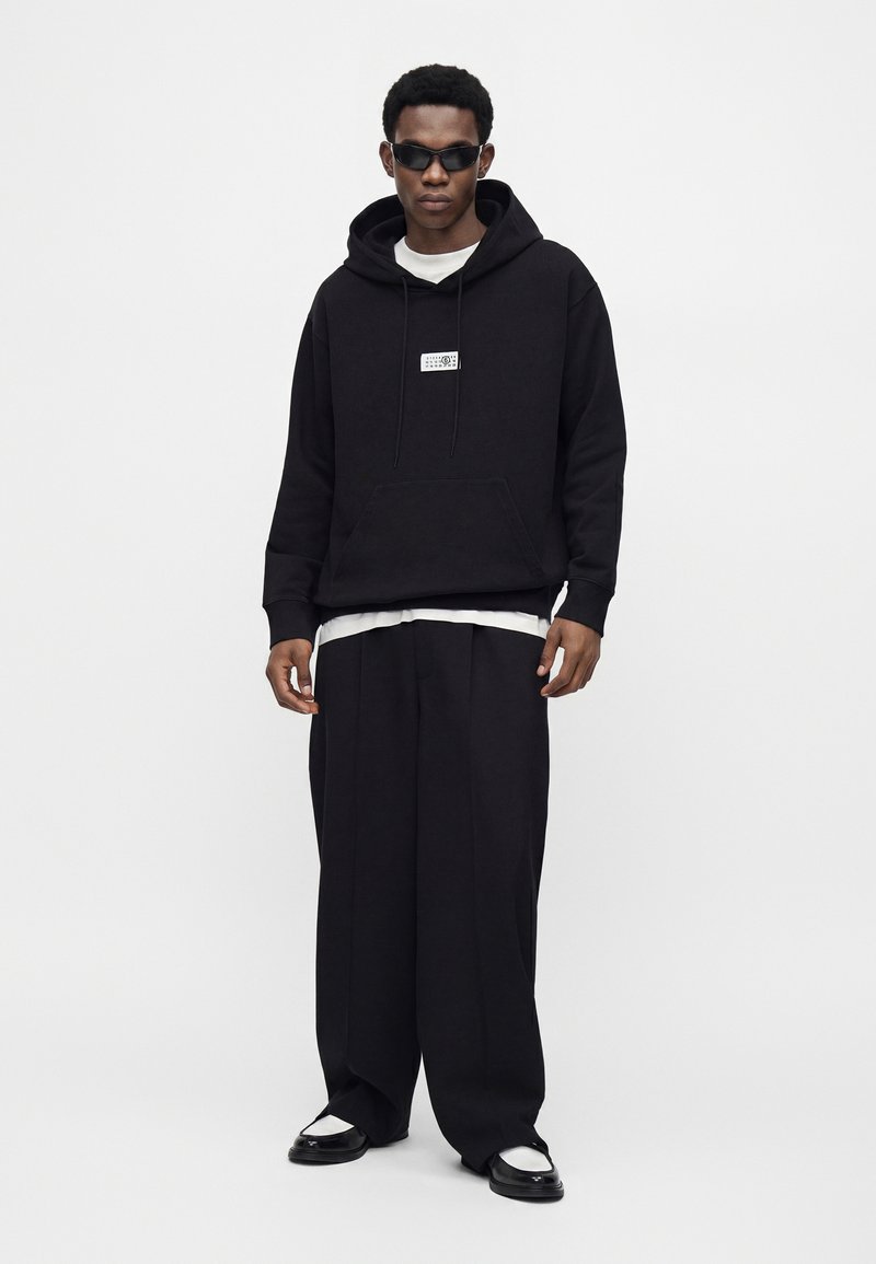 Homme portant un sweat à capuche noir, un pantalon large noir, des baskets blanches et des lunettes de soleil foncées, debout devant un fond blanc.